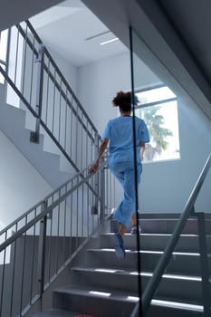 Rear view of mixed race female doctor wearing scrubs running up stairs in hospital. medical professional at work.