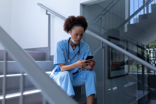 Mixed race female doctor with face mask using smartphone sitting on stairs in hospital. medical professional at work during coronavirus covid 19 pandemic.