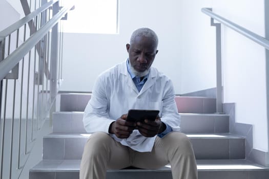 Senior african american male doctor using tablet sitting on stairs in hospital. medical professional at work.