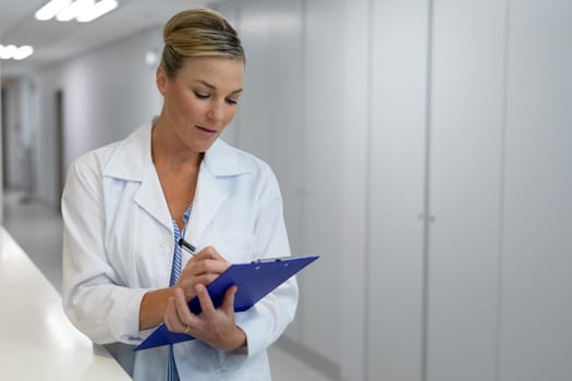 Caucasian female doctor standing in hospital corridor filling medical chart document. medicine, health and healthcare services.