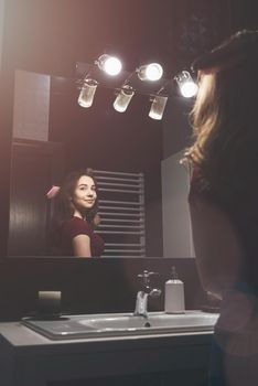 Young woman brushing hair in front of a bathroom mirror. Black tiles on a wall