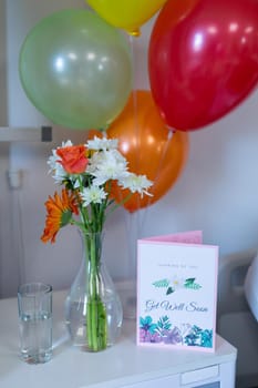 Flowers in vase, get well card and balloons on bedside table in children's ward at a hospital. medicine, health and healthcare services.