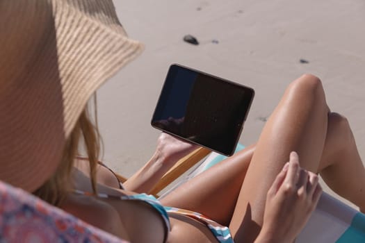 Mid section of caucasian woman using digital tablet while sitting on deck chair at the beach. summer beach holiday concept.
