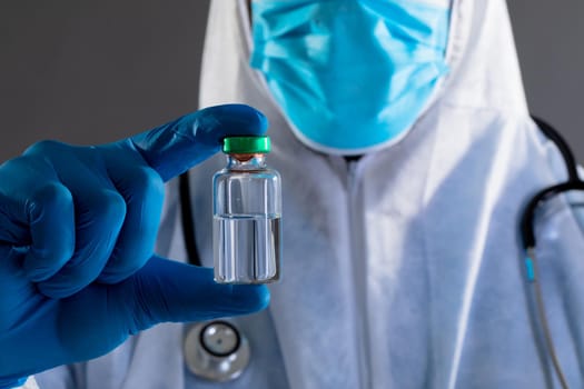 Mid section of health worker wearing protective clothes holding covid-19 vaccine bottle. vaccination for prevention of coronavirus outbreak concept