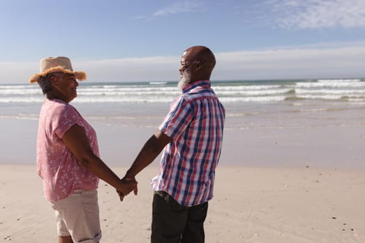 Senior african american couple smiling looking at each other while holding hands on the beach. travel vacation retirement lifestyle concept