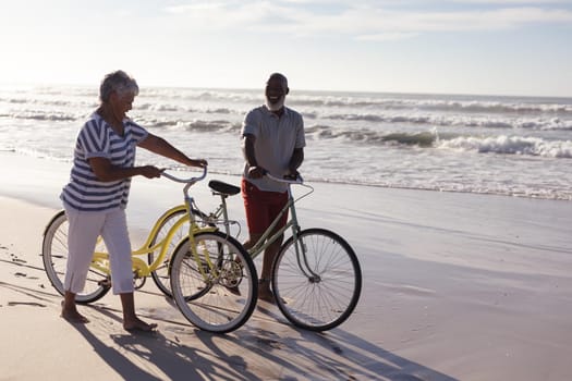 Happy senior african american couple with bicycles walking together on the beach. travel vacation retirement lifestyle concept