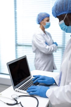 African american male doctor wearing mask and latex gloves using laptop with copy space on screen. medicine, health and healthcare services during coronavirus covid 19 pandemic. .