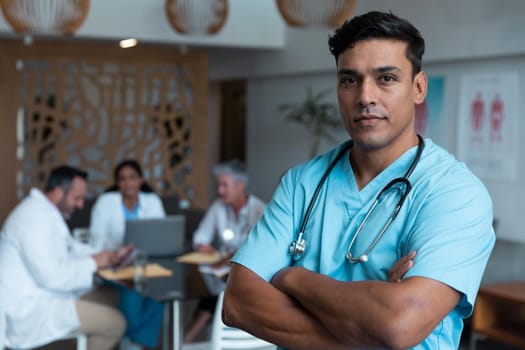 Portrait of mixed race male doctor with arms crossed, colleagues in discussion in the background. medicine, health and healthcare services.
