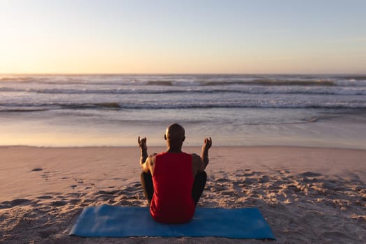 Rear view of senior african american man meditating and practicing yoga while sitting on yoga mat at the beach. fitness yoga and healthy lifestyle concept