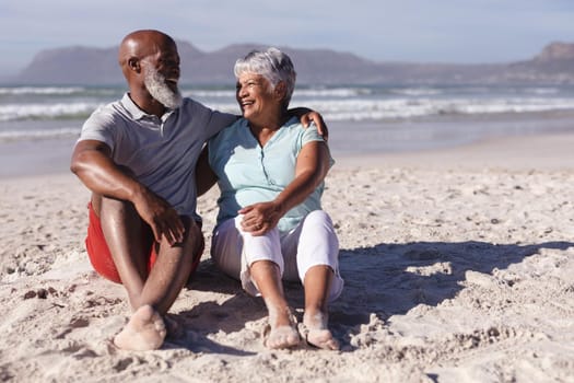 Senior african american couple smiling while looking at each other sitting on the beach. travel vacation retirement lifestyle concept