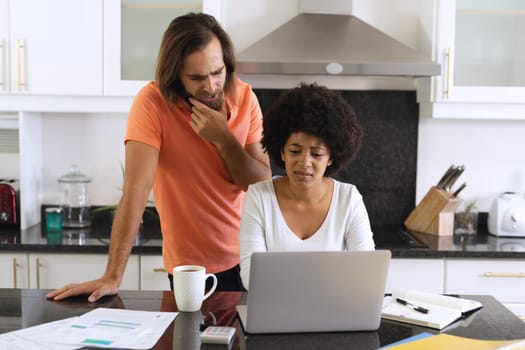 Diverse couple sitting in kitchen using laptop and paying bills. staying at home in isolation during quarantine lockdown.