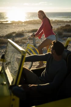 Happy caucasian couple on beach during sunset, woman riding bicycle, man sitting in beach buggy. beach break on summer holiday road trip.