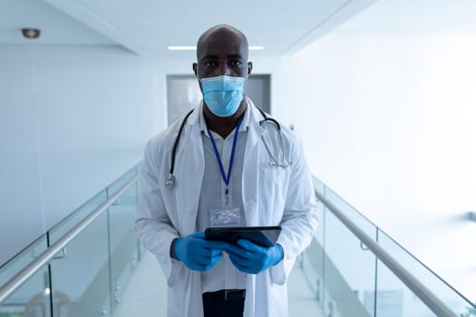 Portrait of african american male doctor in hospital corridor wearing face mask using tablet. medical professional at work during coronavirus covid 19 pandemic.