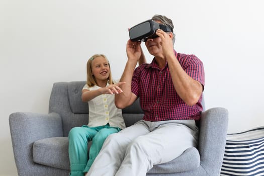 Caucasian granddaughter sitting on couch beside grandfather helping with his vr headset. happy family spending time together at home.