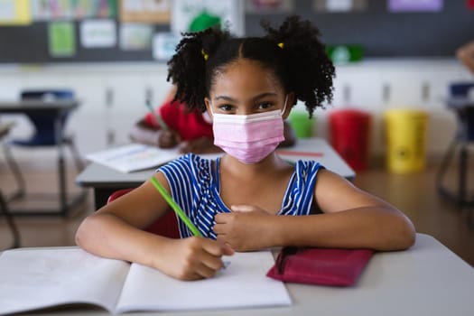 Portrait of african american girl wearing face mask sitting on her desk in the class at school. education back to school health safety during covid19 coronavirus pandemic