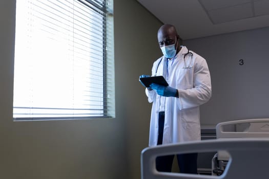 African american male doctor in hospital wearing face mask using tablet. medical professional at work during coronavirus covid 19 pandemic.