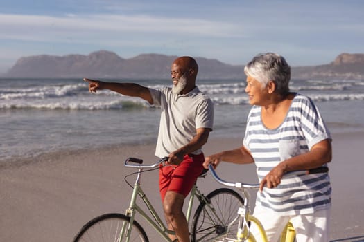 Senior african american couple with bicycles pointing towards a direction at the beach. travel vacation retirement lifestyle concept