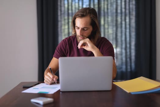 Caucasian man sitting at table working from home, making notes and using laptop. staying at home in isolation during quarantine lockdown.