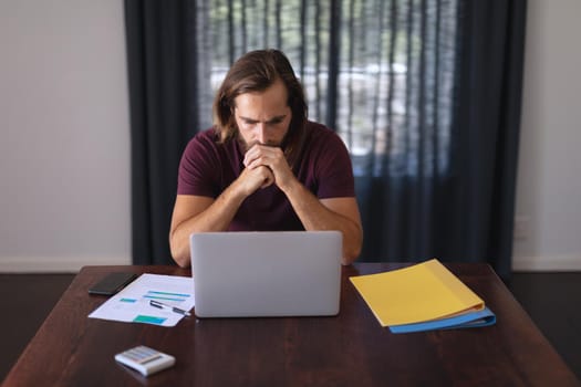 Worried caucasian man sitting at table working from home and looking at laptop screen. staying at home in isolation during quarantine lockdown.