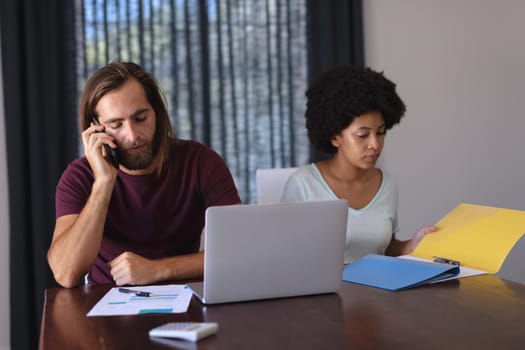 Diverse couple by table working from home, man talking on smartphone, woman checking documents. staying at home in isolation during quarantine lockdown.