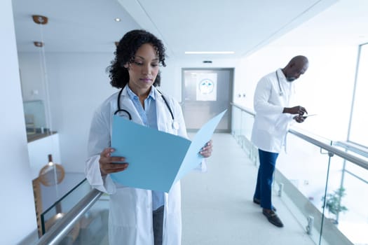 Diverse male and female doctor reading patient file and using digital tablet in hospital corridor. medicine, health and healthcare services.