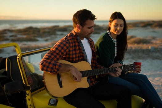 Happy caucasian couple sitting on beach buggy by the sea playing guitar and drinking during sunset. beach break on summer holiday road trip.