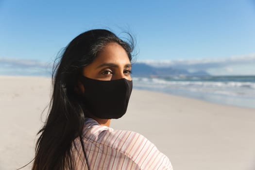 Portrait of mixed race woman on beach holiday wearing face mask. outdoor leisure vacation time by the sea during coronavirus covid 19 pandemic.