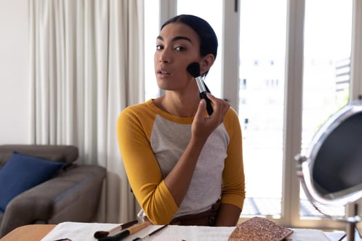 Mixed race gender fluid man putting on foundation powder with makeup brush. staying at home in isolation during quarantine lockdown.