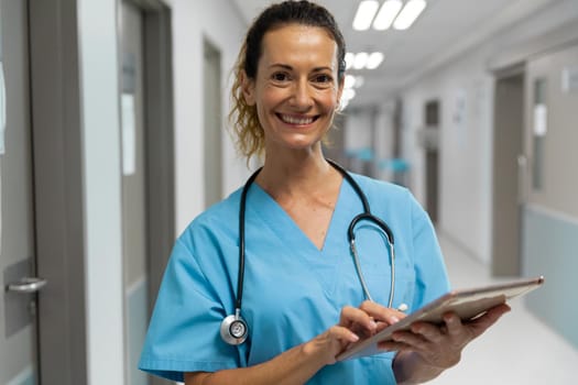 Portrait of mixed race female doctor stading on corridor smiling using laptop. medicine, health and healthcare services.
