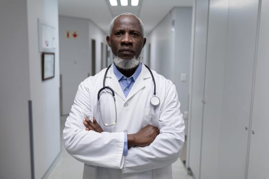Portrait of senior male doctor with stethoscope wearing lab coat, arms crossed in hospital corridor. medical professional at work.