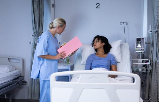 Caucasian female doctor holding file talking with mixed race patient sitting up in hospital bed. medicine, health and healthcare services.