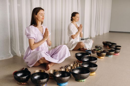 Two women are sitting with Tibetan bowls in the lotus position before a yoga class in the gym.