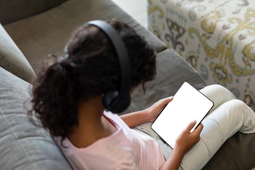 Elevated view of mixed race girl wearing wireless headphones sitting on couch using digital tablet. teenage lifestyle, leisure time, communication and technology.