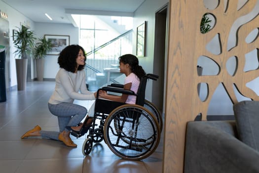 Happy mixed race mother kneeling holding hands talking with daughter in wheelchair in hospital foyer. medicine, health and healthcare services.