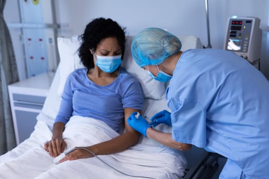 Caucasian female doctor wearing face mask vaccinating african american female patient. medical professional at work during coronavirus covid 19 pandemic.