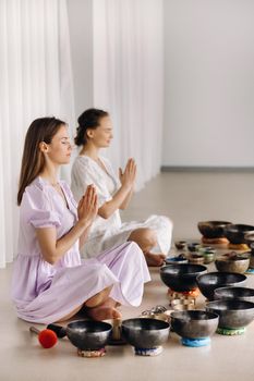 Two women are sitting with Tibetan bowls in the lotus position before a yoga class in the gym.