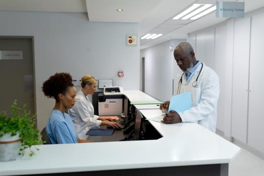 Three diverse male and female doctors in hospital reception using computers looking at documentation. medicine, health and healthcare services.