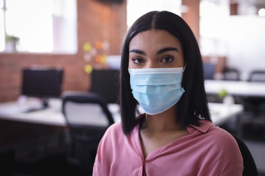 Mixed race businesswoman sitting in office wearing face mask. independent creative design business during covid 19 coronavirus pandemic.