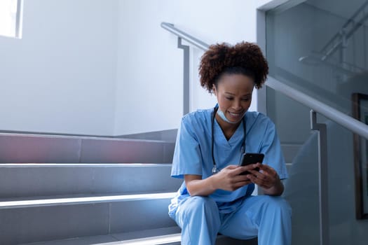 Smiling mixed race female doctor with face mask using smartphone sitting on stairs in hospital. medical professional at work during coronavirus covid 19 pandemic.