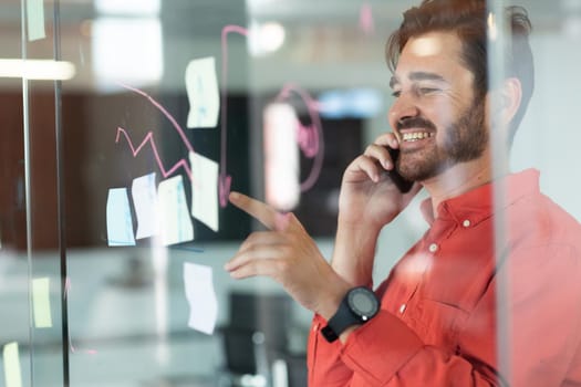 Portrait of caucasian businessman talking on smartphone in front of glass wall with drawings. independent creative design business.