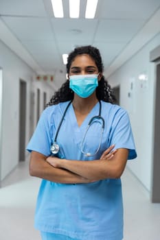 Portrait of mixed race female doctor wearing face mask standing in hospital corridor. medicine, health and healthcare services during coronavirus covid 19 pandemic.