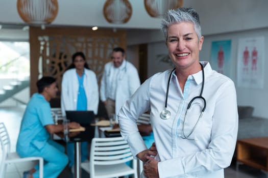 Portrait of smiling caucasian female senior doctor, with colleagues in discussion in the background. medicine, health and healthcare services.