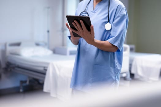 Midsection of caucasian female doctor in hospital wearing scrubs and stethoscope using tablet. medical professional at work and health services.