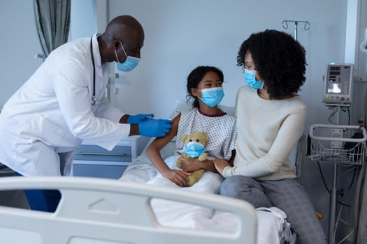 Male doctor giving covid vaccination to sick girl in hospital bed with mother, all in face masks. medicine, health and healthcare services during coronavirus covid 19 pandemic.
