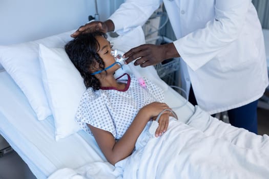 Midsection of male doctor putting oxygen mask ventilator on sick mixed race girl in hospital bed. medicine, health and healthcare services during coronavirus covid 19 pandemic.