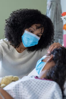 Mixed race mother and sick daughter in face masks in hospital, girl holding teddy bear. medicine, health and healthcare services during covid 19 coronavirus pandemic.