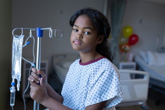 Portrait of bored sick mixed race girl standing in hospital ward with iv drip bag on stand. medicine, health and healthcare services.