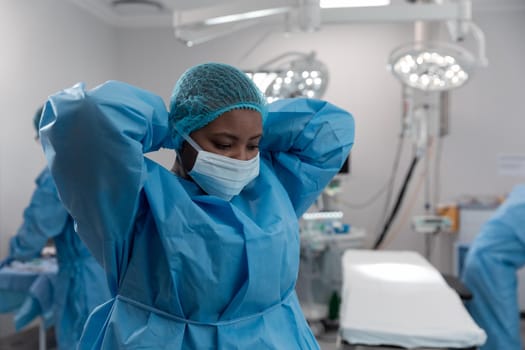 Portrait of mixed race female surgeon in operating theatre putting on face mask. medicine, health and healthcare services during coronavirus covid 19 pandemic.