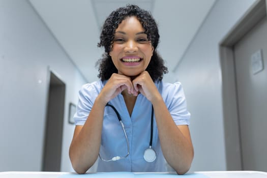Mixed race female doctor at desk leaning chin on hands and smiling during video call consultation. telemedicine, online healthcare services during quarantine lockdown.