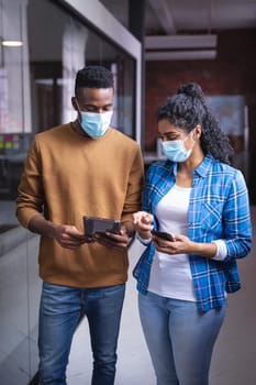 Diverse male and female colleagues at work discussing and looking at tablet wearing face masks. independent creative design business during covid 19 coronavirus pandemic.